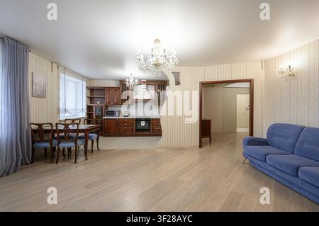 heller, offener Wohnbereich mit einer Küche. Enthält ein hölzernes Esszimmer-Set, ein blaues Sofa und dunkle Holzküchenkästen. Ein Kronleuchter hängt in der Mitte Stockfoto
