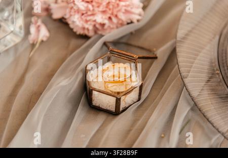 Elegante Goldhochzeitsringe, die in einer geometrischen Glasringbox mit dekorativem Salz präsentiert werden. Romantische Hochzeitstischdekoration mit weichem Stoff, rosa Nelke und stilvollen Details. Nahaufnahme, die Liebe, Ehe und Hingabe symbolisiert. Stockfoto