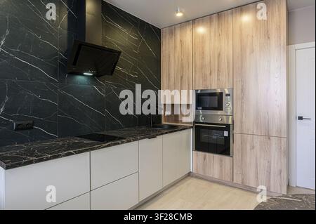 Moderne Küche mit schwarzer Marmor-Rückwand und Arbeitsplatten. Helle Holzschränke integrieren Edelstahlgeräte. Sauberes, minimalistisches Design Stockfoto
