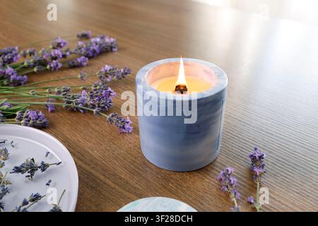 Brennende Kerze in blauem Keramikglas, Nahaufnahme, hölzerner Tisch als Hintergrund. Lilafarbener Lavendelblütenstrauß, Plattenseitenansicht, selektive Fokussierung. Aromatherapie-Spa Stockfoto