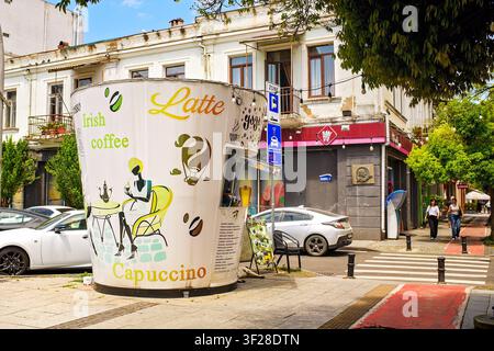 Batumi, Georgien – 9. Juni 2026: Kaffeebude in Form einer riesigen Tasse auf dem Bürgersteig neben dem Zebrastreifen und historischen Gebäuden, Menschen gehen auf der Straße. Stockfoto