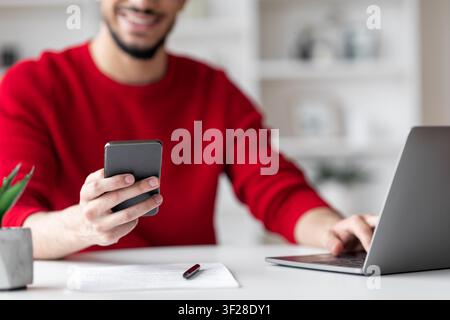 Lächelnder Mann hält Smartphone und arbeitet am Laptop im Homeoffice, Business-Konzept Stockfoto
