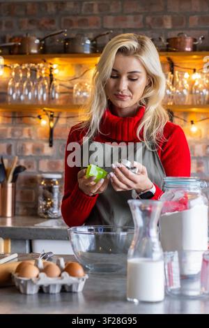 Bäckerin in rotem Pullover und grauer Schürze, die Messbecher in der Küche mit Eiern, Mehl, Milch hält. Gemütlich, rustikal, warm, Backen, Kupfer, Glaswaren, Vorratskammer Stockfoto