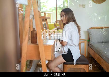 Junge Frau führt ihre Haargestaltungsroutine mit einem Glätteisen in einem gemütlichen modernen Schlafzimmerinneren durch. Stockfoto