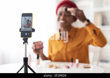 Lächelnde schwarze Frau, die vor einem Smartphone auf einem Stativ Make-up aufträgt, Konzept für häusliche Hautpflege Stockfoto