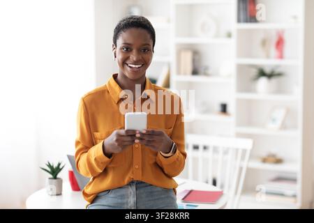 Lächelnde schwarze Frau, die zu Hause ein Smartphone hält, Geschäftskonzept Stockfoto