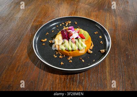 Gesundes Obstdessert auf einem schwarzen Teller mit Avocado, Banane, Drachenfrucht, Erdbeere, Kiwi und Granola auf einem Holztisch in einem Café-Esszimmer-Set Stockfoto