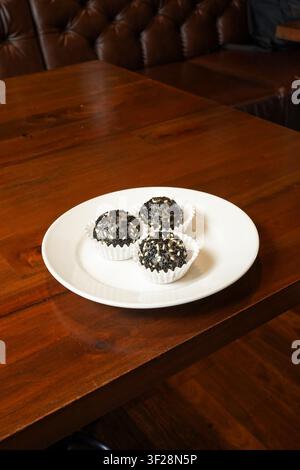 Drei schwarze Sesam-Dessertbällchen, die auf einem weißen Teller auf einem Holztisch serviert werden. Süßes asiatisches Dessert mit Sesamüberzug in einer lockeren Restaurantatmosphäre. Stockfoto