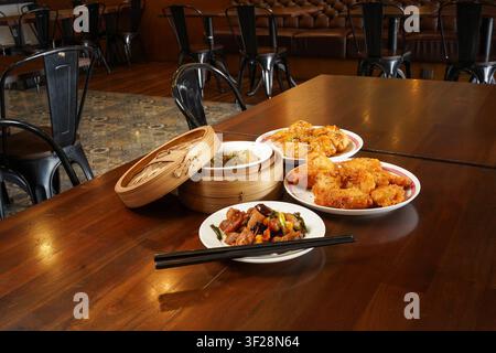 Vielzahl asiatischer Gerichte wie Dim Sum, gebratenes Huhn, Garnelen und Wok-Gericht, serviert auf einem Restauranttisch mit Milchtee in einer zwanglosen Essensumgebung. Stockfoto
