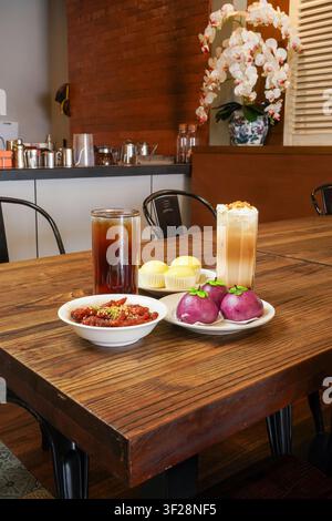 Verschiedene asiatische Desserts und Getränke, darunter Vanillekuchenbrötchen, lila fruchtförmige Brötchen, Eiskaffee und Tee, serviert auf einem Holztisch in einem gemütlichen Restaurant Stockfoto