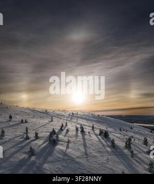 Luftbild des Yllas-Berges im Winter mit schneebedeckten Hängen, vereinzelten Kiefern, langen Schatten und einer hellen Sonne mit Sonne-Hunden am Himmel in Kol Stockfoto