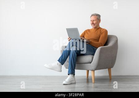 Zufriedener älterer kaukasischer Mann lächelt, während er in einem gemütlichen Wohnzimmer einen Laptop benutzt, Seniorenlebensstil-Konzept Stockfoto
