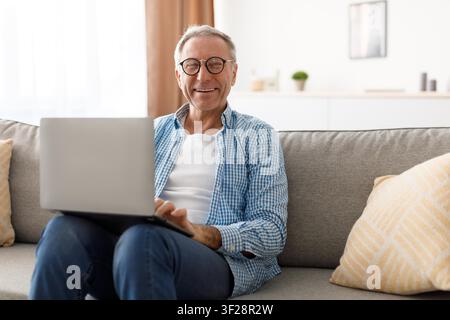 Zufriedener älterer kaukasischer Mann, der zu Hause auf dem Sofa einen Laptop benutzt, Geschäftskonzept Stockfoto
