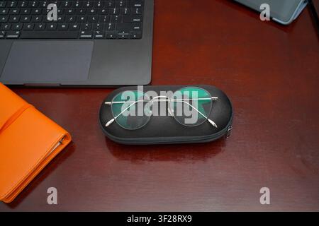 Runde Brille, die auf einem Etui neben einem Laptop und einem orangefarbenen Notizbuch auf einem Holzschreibtisch liegt, repräsentiert Arbeitsplatzutensilien, Produktivität und moderne o Stockfoto