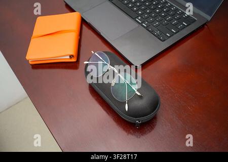 Runde Brille, die auf einem Etui neben einem Laptop und einem orangefarbenen Notizbuch auf einem Holzschreibtisch liegt, repräsentiert Arbeitsplatzutensilien, Produktivität und moderne o Stockfoto