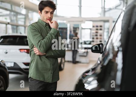 Nachdenklicher junger kaukasischer Mann, der den Kauf eines Autos in der Ausstellungshalle überlegt, Geschäftskonzept Stockfoto