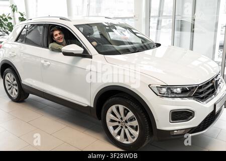 Glücklicher junger Mann lächelt in weißem SUV-Auto im Autohaus, begeistert, Fahrzeug zu kaufen Stockfoto