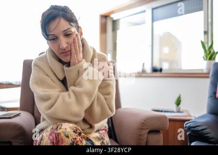 Junge Frau sitzt auf einem Sessel, hält ihr Gesicht und fühlt sich traurig und deprimiert Stockfoto