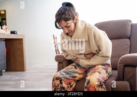 Frau, die starke Magenschmerzen und Krämpfe verspürt, während sie zu Hause auf einem Stuhl sitzt Stockfoto