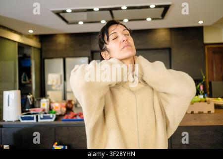 Frau mit Nackenschmerzen und Verspannungen reibt ihre Nackenmuskeln in einer modernen Küche Stockfoto