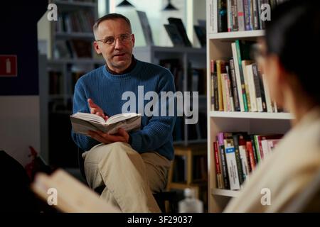 Mittealter Mann kaukasischer Abstammung liest Buch und führt Diskussion in der Bibliothek, unterstützt erwachsenes Lernen und Gruppenbildung. Nützlich für Alphabetisierungsprogramme, Workshops, Buchclubs Stockfoto