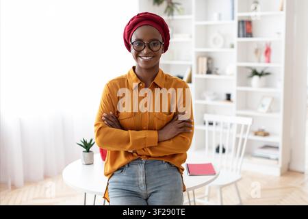 Lächelnde schwarze Frau mit rotem Kopftuch und Brille steht in einem hellen modernen Büro Stockfoto