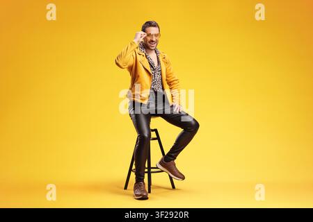 Stylischer Mann in einer gelben Jacke, der auf einem Hocker isoliert auf gelbem Hintergrund sitzt Stockfoto