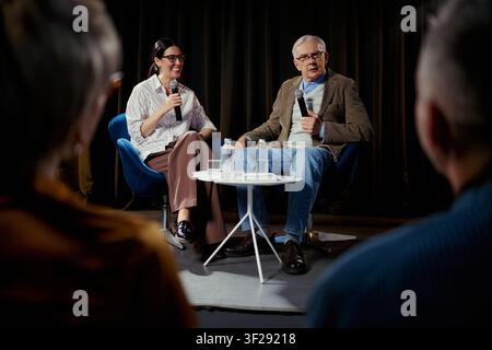 Breite Aufnahme eines älteren kaukasischen Mannes und einer jungen Frau, die während einer Podiumsdiskussion in Mikrofone sprechen und Expertise sowie Führungskenntnisse teilen. Für Konferenzmarketing und Unternehmenskommunikation Stockfoto
