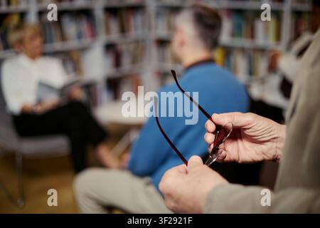 Seniorin führt Gruppendiskussion in der Bibliothek, mittlerer Mann hält eine Brille während er während einer Beratungssitzung zuhört. Geeignet für psychische Gesundheit, Selbsthilfegruppe, Therapie-Marketing Stockfoto