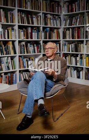 Vertikale Aufnahme eines älteren kaukasischen Mannes, der ein Buch liest, während er vor Bücherregalen sitzt, was lebenslanges Lernen und Wissen suggeriert. Nützlich für Bildung, Verlagswesen, Ruhestandsinhalte Stockfoto
