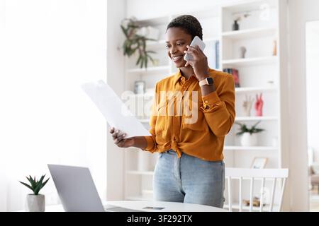 Lächelnde schwarze Frau telefoniert, während sie zu Hause im Büro ein Dokument hält, Geschäftskonzept Stockfoto