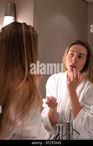 Frau trägt Lippenbalsam auf, während sie im Badezimmerspiegel während ihrer morgendlichen Schönheitsroutine in einem Luxushotelzimmer schaut Stockfoto