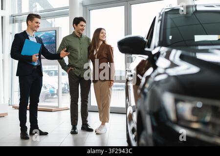Junges Paar berät sich mit einem männlichen Autoverkäufer im Ausstellungsraum, Autokaufprozess Stockfoto