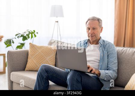 Senior weißer Mann arbeitet an einem Laptop zu Hause, konzentriert im modernen Wohnzimmer Stockfoto