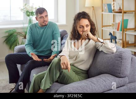 Junger Ehemann spricht mit seiner unglücklichen, verärgerten Frau, die zu Hause auf dem Sofa sitzt. Stockfoto