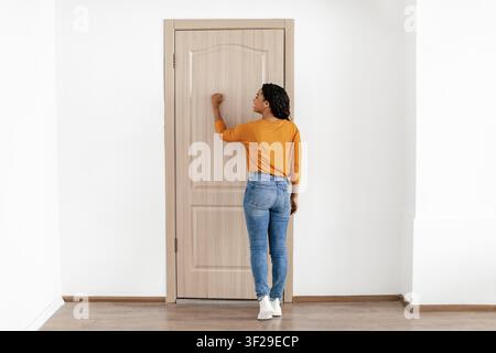 Schwarze Frau klopft an hölzerne Tür in häuslicher Umgebung, nicht zu erkennen. Zuhause-Konzept Stockfoto