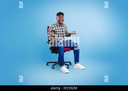Lächelnder schwarzer Mann, der eine Handgeste verwendet, während er auf einem Bürostuhl sitzt, Studioneinstellung Stockfoto