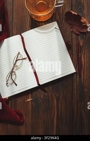 Gemütlicher Herbst-Flat-Lay mit einem offenen linierten Notizbuch, einer Lesebrille, einem roten Stift, einem warmen Getränk, einem Ahornblatt und einem gestrickten Schal auf dunklem Holz Stockfoto