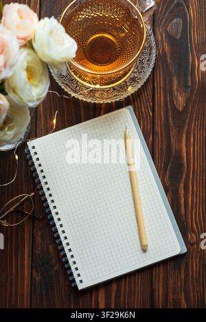 Offenes Spiralnotizbuch mit einem Stift, einer Tasse Tee, einem Rosenstrauß, einer Lesebrille und Lichterketten, die eine gemütliche, inspirierende Atmosphäre auf einem dunklen Hintergrund schaffen Stockfoto