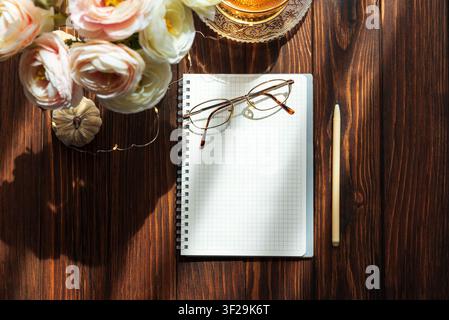Holzschreibtisch-Arbeitsplatz mit einem offenen Spiralnotizbuch mit Millpapier und Lesebrille, einem Stift, einer Tasse und Blumen, alles beleuchtet von warmem Morgenlicht Stockfoto