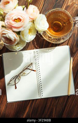 Offenes Gitter-Notizbuch mit einer Lesebrille und einem Stift, der auf einem dunklen Holztisch neben einer Tasse heißem Tee und einem Strauß Ranunkulusblumen unter warmem Licht liegt Stockfoto