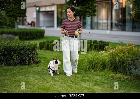 Japanische Frau führt ihren französischen Bulldoggen an der Leine im Stadtpark spazieren Stockfoto