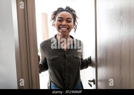 Lächelnde schwarze Frau öffnet die Tür mit herzlicher Begrüßung zu Hause, Glücksbegriff Stockfoto