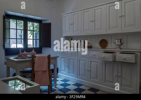 Antike Küche im Castillo Gala Dalí in Púbol, Girona, Spanien, mit weißen Holzschränken, einem karierten Fliesenboden und Blick durch ein offenes Fenster. Stockfoto