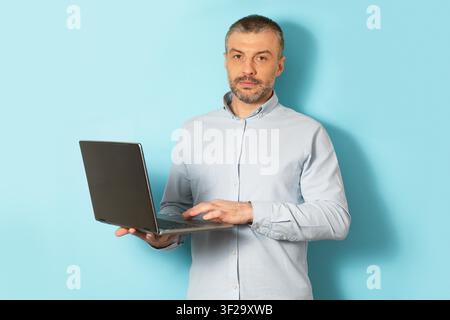 Ernster Kaukasier verwendet einen Laptop, steht vor hellblauer Kulisse, Technologiekonzept Stockfoto