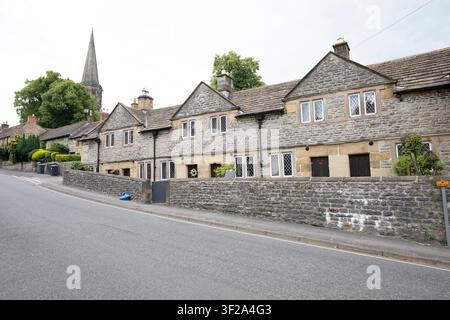 Die historischen St. John's Hospital Almshouses hinter einer Steinmauer auf einem steilen Hügel, mit dem Turm der All Saints Kirche in Bakewell, Derbyshire, UK. Stockfoto