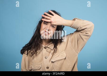 Frustrierte junge Frau mit dunklen Haaren, die die Stirn hält, blauer Hintergrund, Stresskonzept Stockfoto
