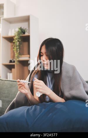 kranke asiatische junge Frau, die in ein Papiertaschentuch niest, während sie mit einer Decke zugedeckt auf dem Sofa im Wohnzimmer sitzt Stockfoto