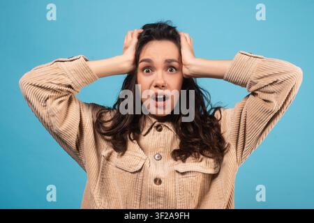 Gestresste junge Frau mit dunklen Haaren, die schockiert aussieht, den Kopf hält, blauer Hintergrund Stockfoto