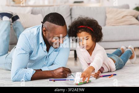 Kreative Lockdown-Aktivitäten. Afroamerikanisches Mädchen und Großvater zeichnen mit Markierungen auf dem Boden im Wohnzimmer Stockfoto